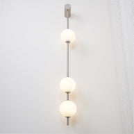 Wall lamp with a vertical row of white ball shades on a slim nickel-coloured rod lit beside a lace curtain.