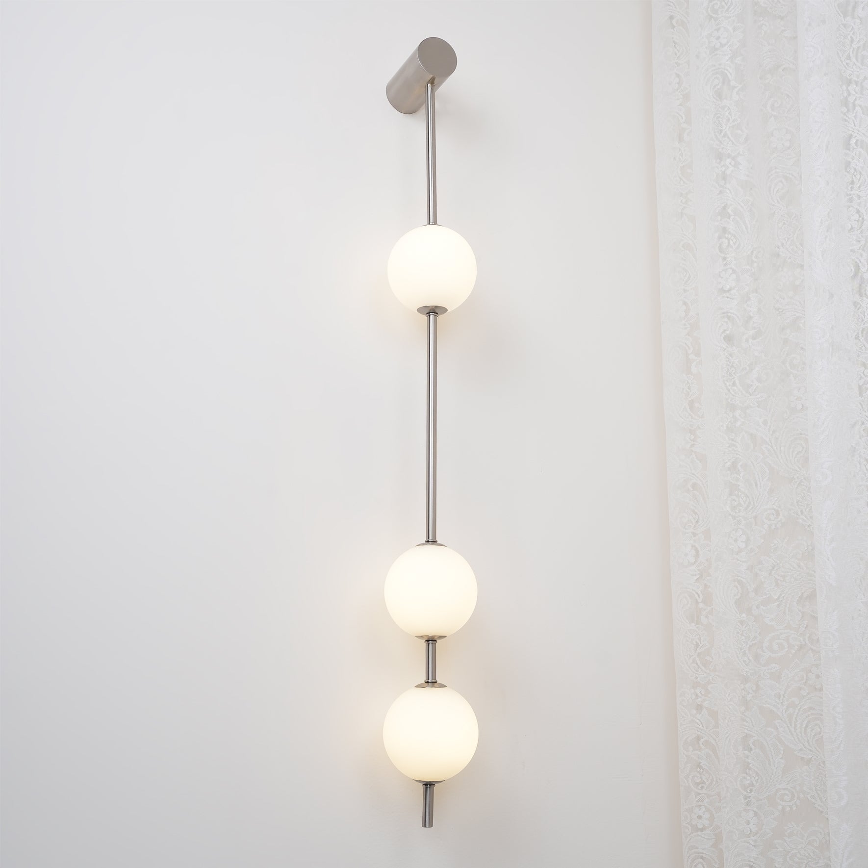 Wall lamp with a vertical row of white ball shades on a slim nickel-coloured rod lit on a white wall.