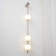 Wall lamp with a vertical row of white ball shades on a slim nickel-coloured rod lit on a white wall.