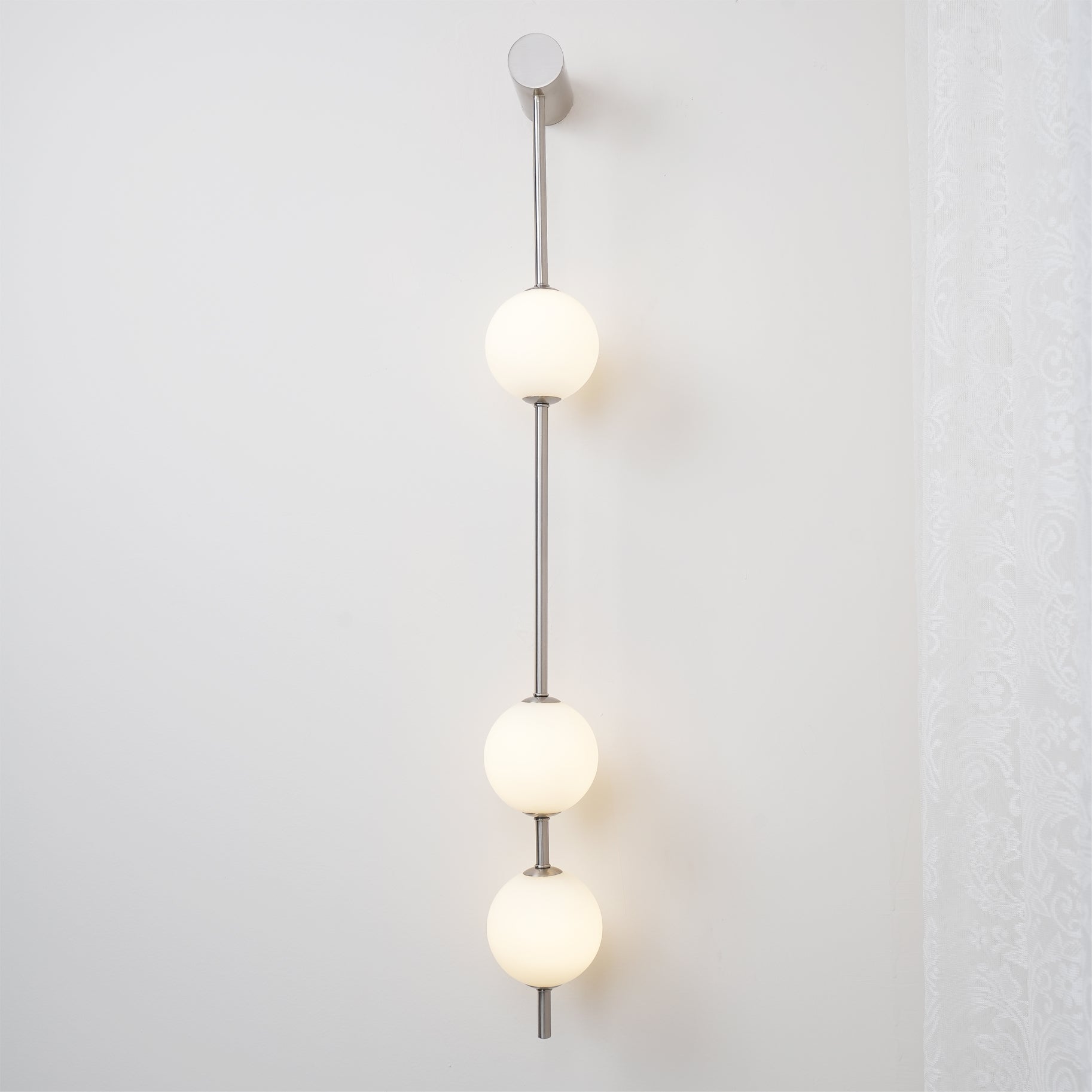 Wall lamp with a vertical row of white ball shades on a slim nickel-coloured rod lit beside a lace curtain.