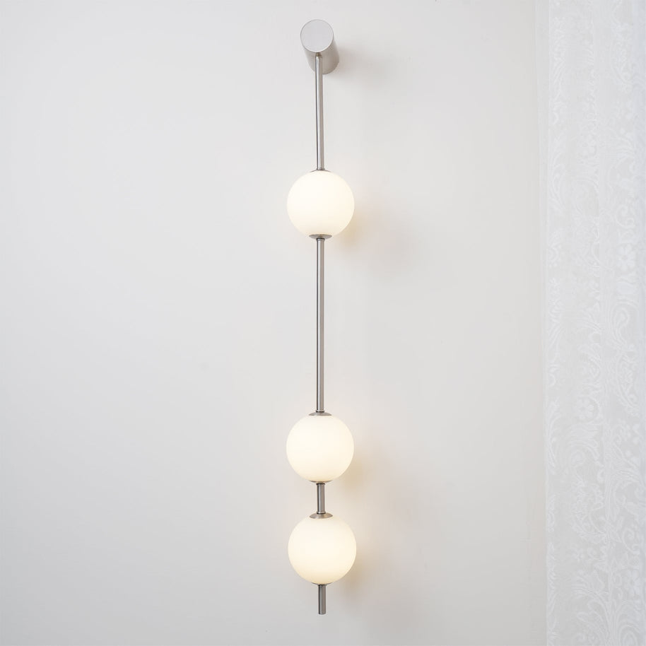 Wall lamp with a vertical row of white ball shades on a slim nickel-coloured rod lit beside a lace curtain.