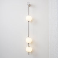 Wall lamp with a vertical row of white ball shades on a slim nickel-coloured rod lit beside a lace curtain.