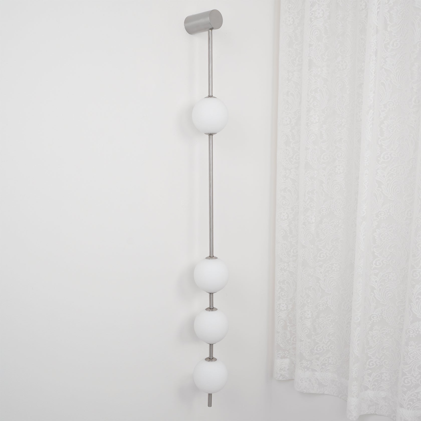 Wall lamp with a vertical row of white ball shades on a slim nickel-coloured rod, unlit, beside a lace curtain.