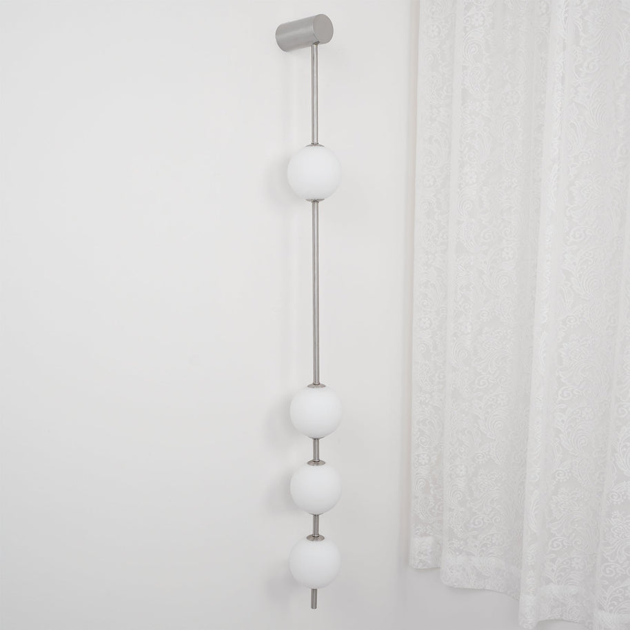 Wall lamp with a vertical row of white ball shades on a slim nickel-coloured rod, unlit, beside a lace curtain.