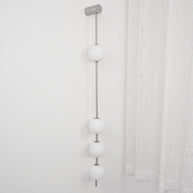 Wall lamp with a vertical row of white ball shades on a slim nickel-coloured rod, unlit, beside a lace curtain.