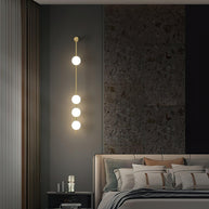 Wall lamp with a vertical row of white ball shades on a slim brass-coloured rod lit on a slatted wall beside a bed.