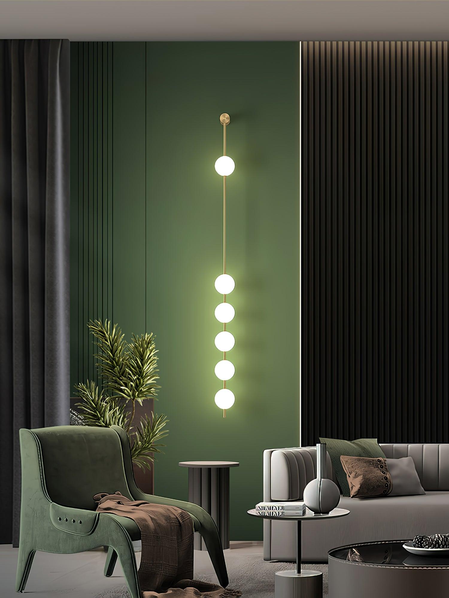 Wall lamp with a vertical row of white ball shades on a slim brass-coloured rod lit against a green wall panel.