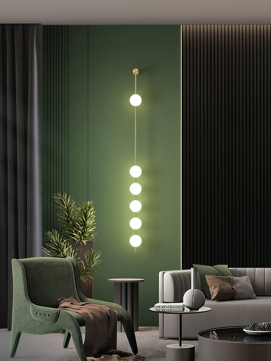 Wall lamp with a vertical row of white ball shades on a slim brass-coloured rod lit against a green wall panel.