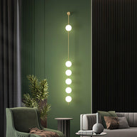 Wall lamp with a vertical row of white ball shades on a slim brass-coloured rod lit against a green wall panel.