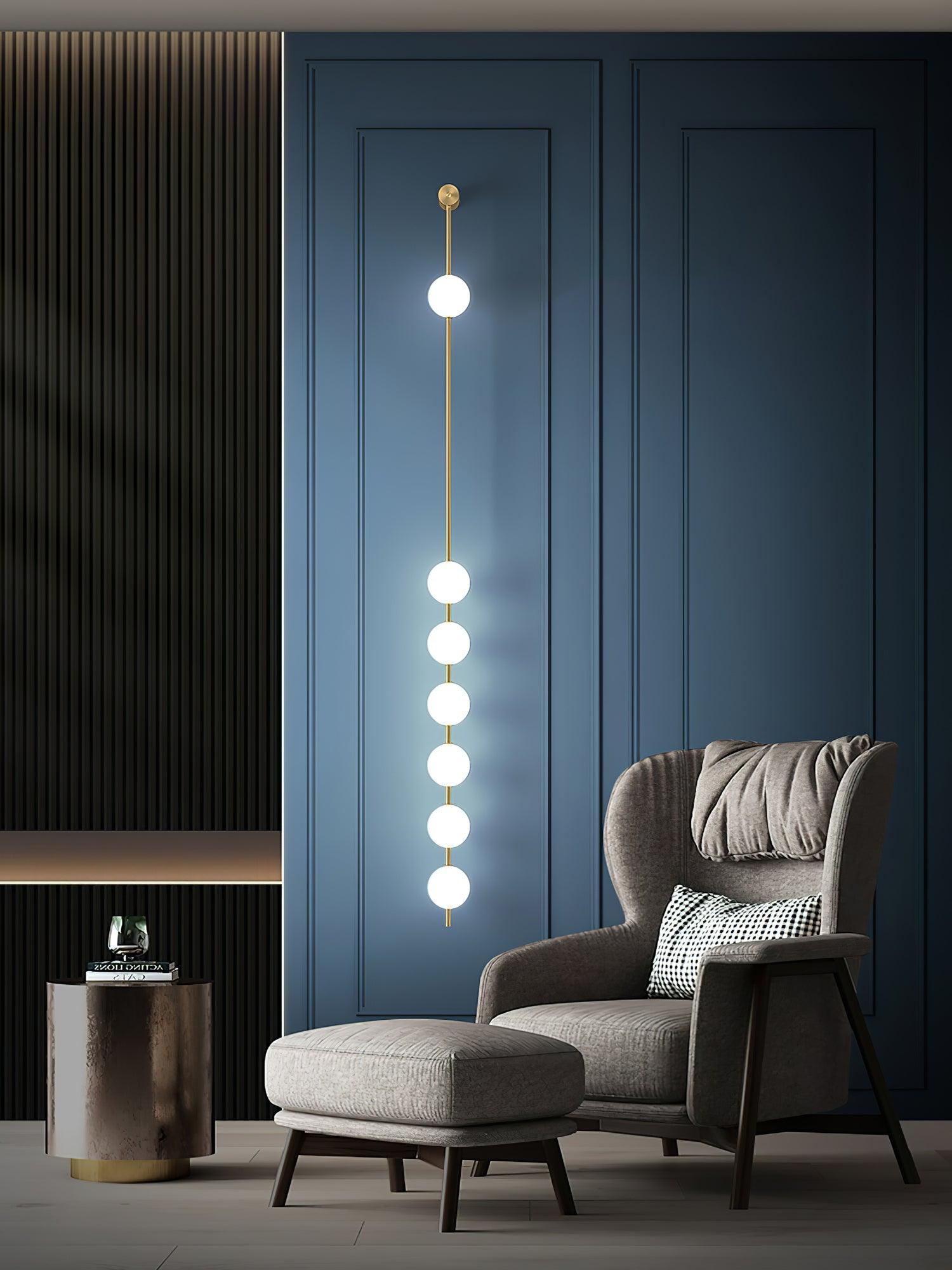 Wall lamp with a vertical row of white ball shades on a slim brass-coloured rod lit on a blue panelled wall beside an armchair.