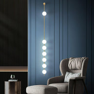 Wall lamp with a vertical row of white ball shades on a slim brass-coloured rod lit on a blue panelled wall beside an armchair.