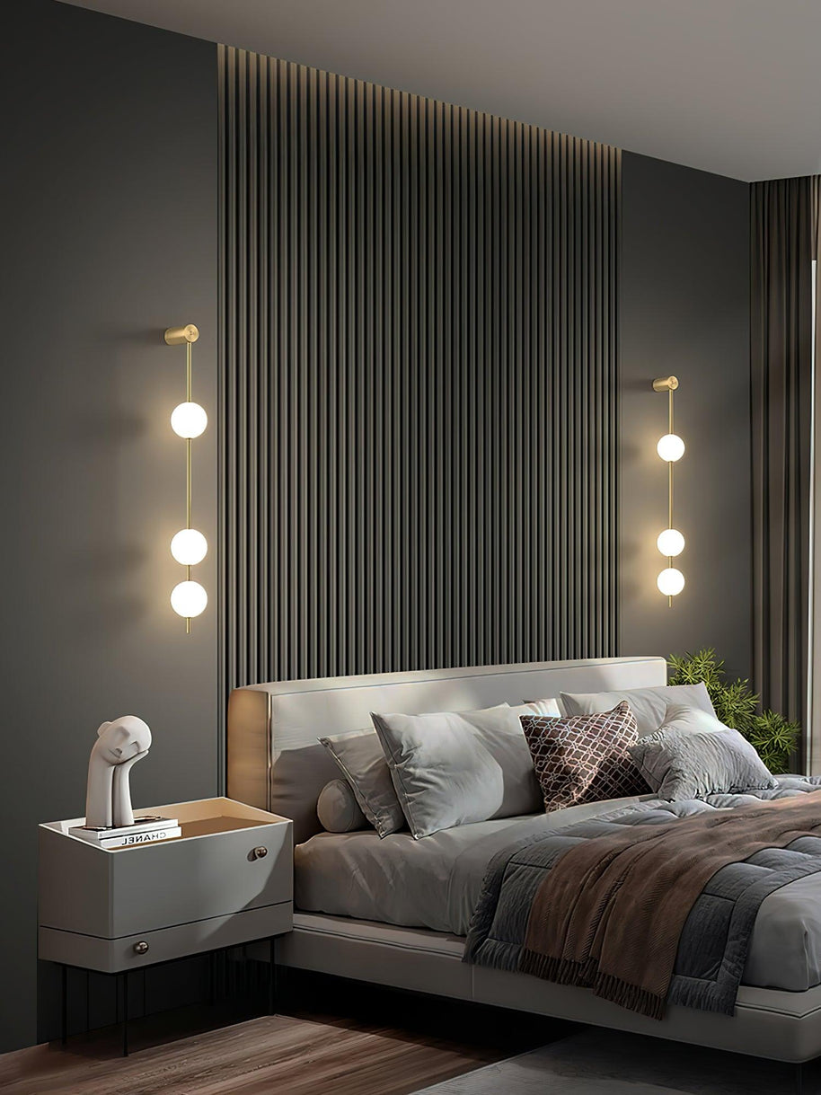 Two wall lamps with vertical rows of white ball shades on slim brass-coloured rods, lit on either side of a bed against a slatted wall.
