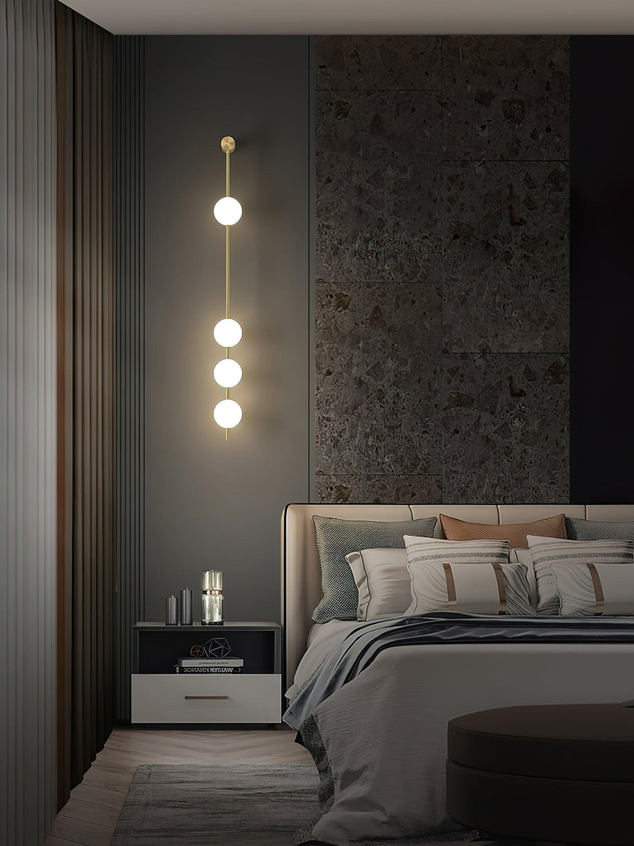 Wall lamp with a vertical row of white ball shades on a slim brass-coloured rod lit on a slatted wall in a bedroom.