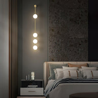 Wall lamp with a vertical row of white ball shades on a slim brass-coloured rod lit on a slatted wall in a bedroom.