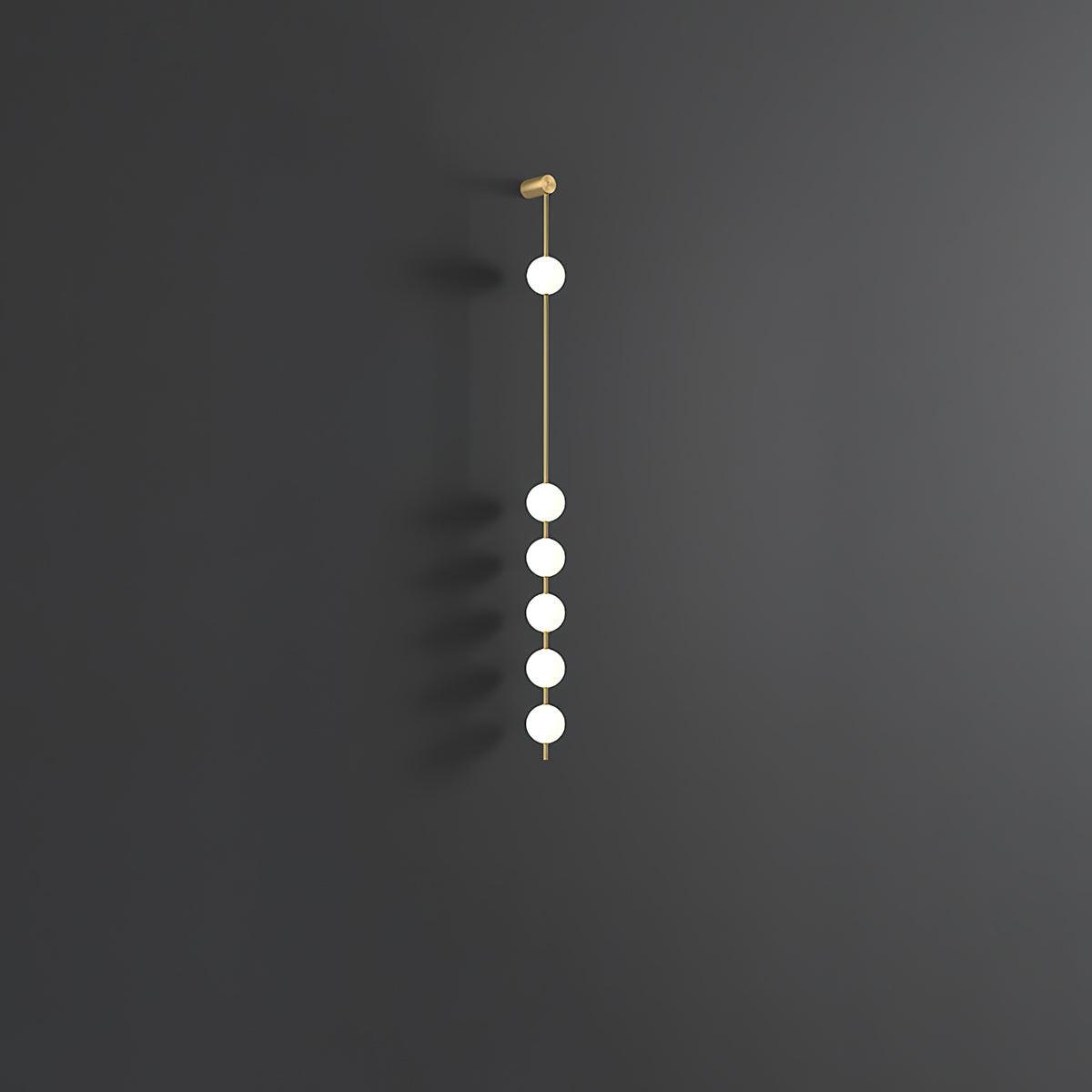 Wall lamp with a vertical row of white ball shades on a slim brass-coloured rod, unlit, on a grey wall.
