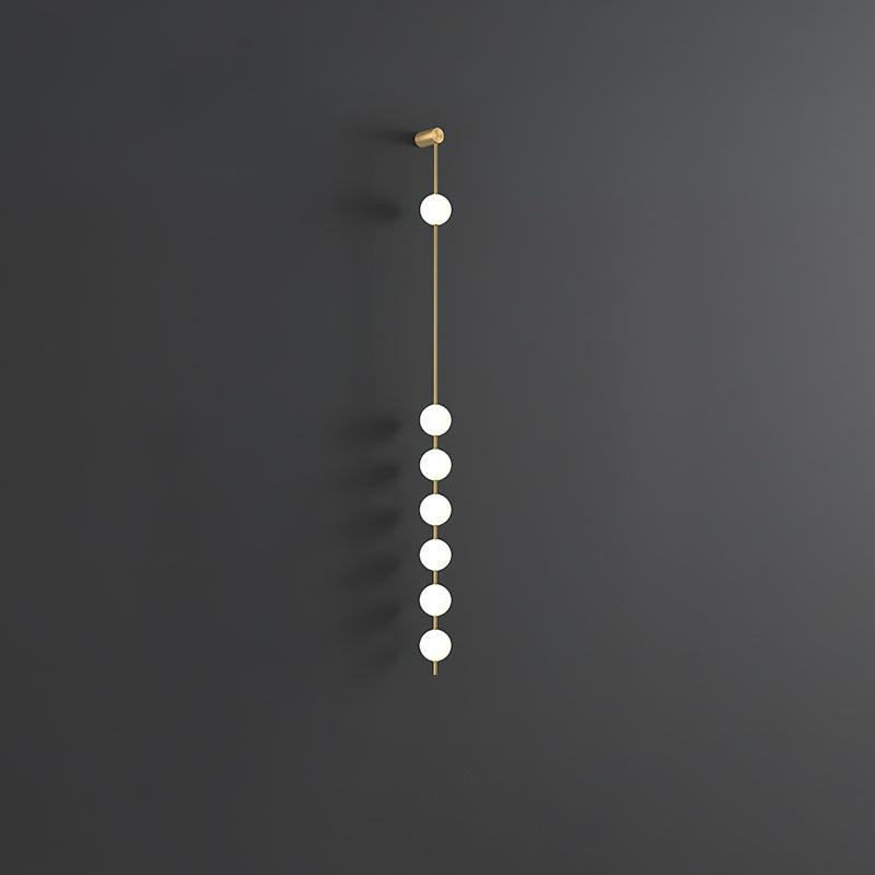 Wall lamp with a vertical row of white ball shades on a slim brass-coloured rod, unlit, on a grey wall, with a longer row of shades.