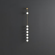 Wall lamp with a vertical row of white ball shades on a slim brass-coloured rod, unlit, on a grey wall, with a longer row of shades.