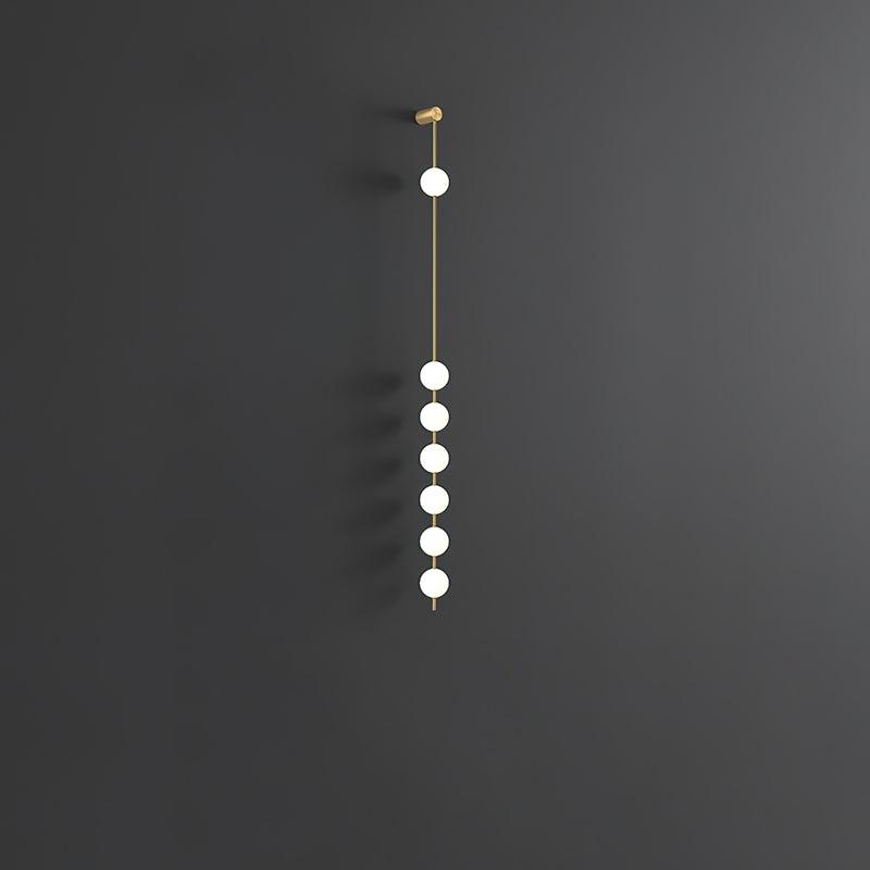 Wall lamp with a vertical row of white ball shades on a slim brass-coloured rod, unlit, on a grey wall.