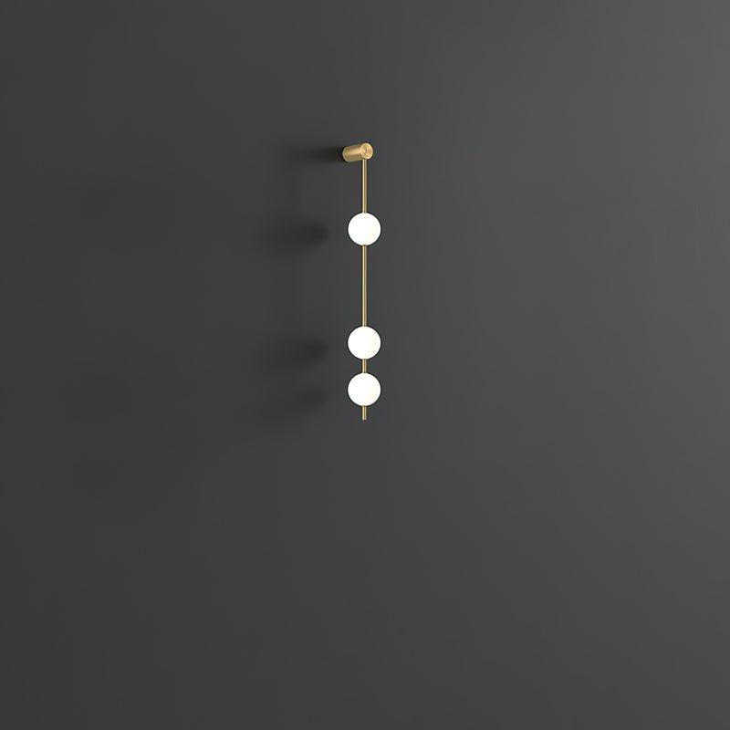 Wall lamp with a vertical row of white ball shades on a slim brass-coloured rod, unlit, on a grey wall, with a short row of shades.