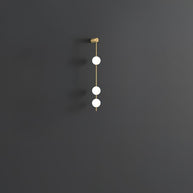 Wall lamp with a vertical row of white ball shades on a slim brass-coloured rod, unlit, on a grey wall, with a short row of shades.