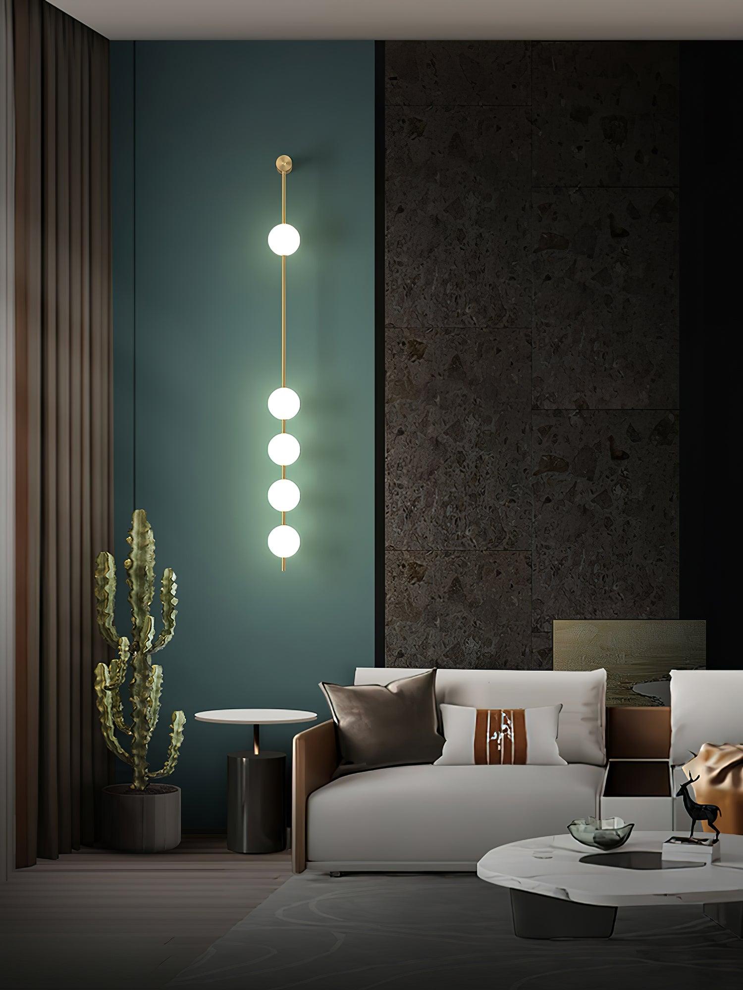 Wall lamp with a vertical row of white ball shades on a slim brass-coloured rod lit against a teal wall panel.