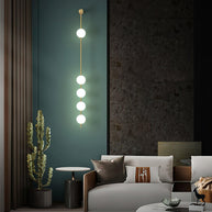 Wall lamp with a vertical row of white ball shades on a slim brass-coloured rod lit against a teal wall panel.