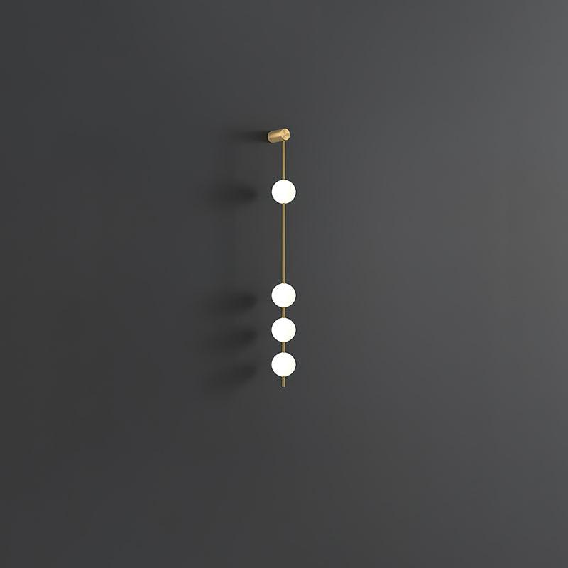 Wall lamp with a vertical row of white ball shades on a slim brass-coloured rod, unlit, on a grey wall.