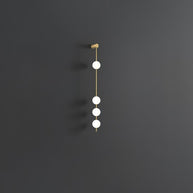 Wall lamp with a vertical row of white ball shades on a slim brass-coloured rod, unlit, on a grey wall.