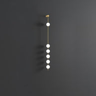 Wall lamp with a vertical row of white ball shades on a slim brass-coloured rod, unlit, on a grey wall.