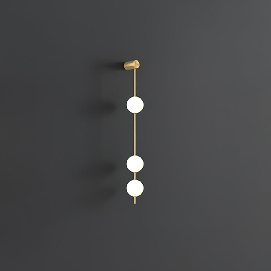 Wall lamp with a vertical row of white ball shades on a slim brass-coloured rod, unlit, on a grey wall, with a short row of shades.