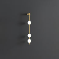 Wall lamp with a vertical row of white ball shades on a slim brass-coloured rod, unlit, on a grey wall, with a short row of shades.