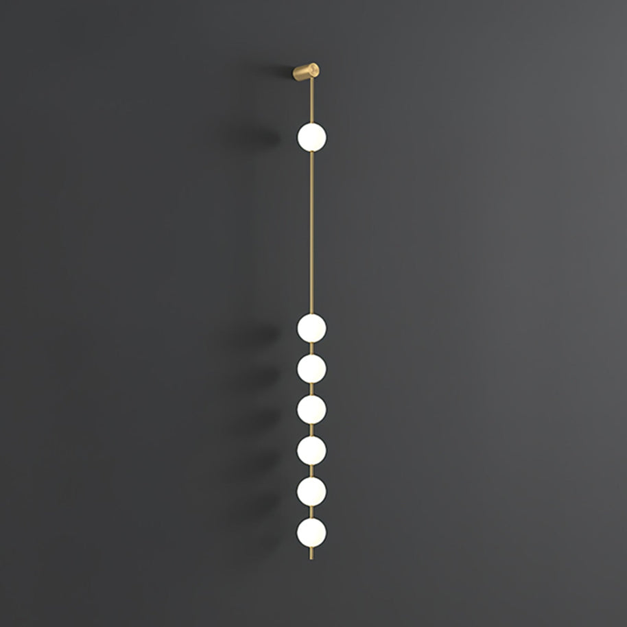 Wall lamp with a vertical row of white ball shades on a slim brass-coloured rod, unlit, on a grey wall, with a longer row of shades.