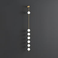 Wall lamp with a vertical row of white ball shades on a slim brass-coloured rod, unlit, on a grey wall, with a longer row of shades.