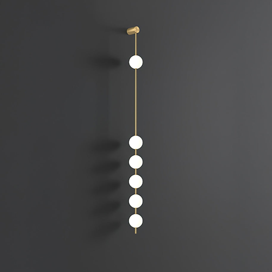 Wall lamp with a vertical row of white ball shades on a slim brass-coloured rod, unlit, on a grey wall.