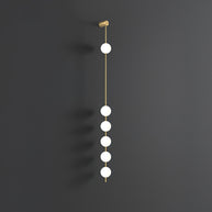 Wall lamp with a vertical row of white ball shades on a slim brass-coloured rod, unlit, on a grey wall.