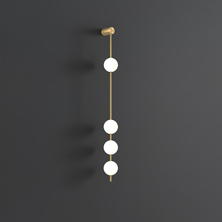 Wall lamp with a vertical row of white ball shades on a slim brass-coloured rod, unlit, on a grey wall.