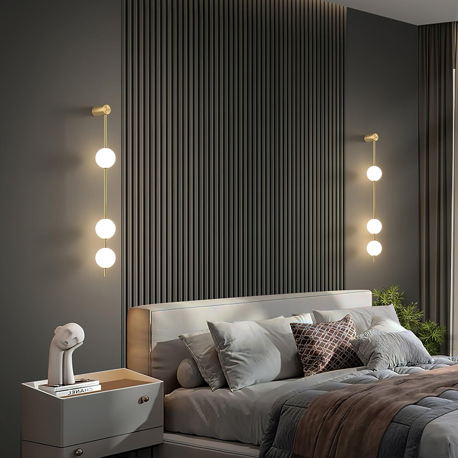 Two wall lamps with vertical rows of white ball shades on slim brass-coloured rods, lit on either side of a bed against a dark slatted wall.