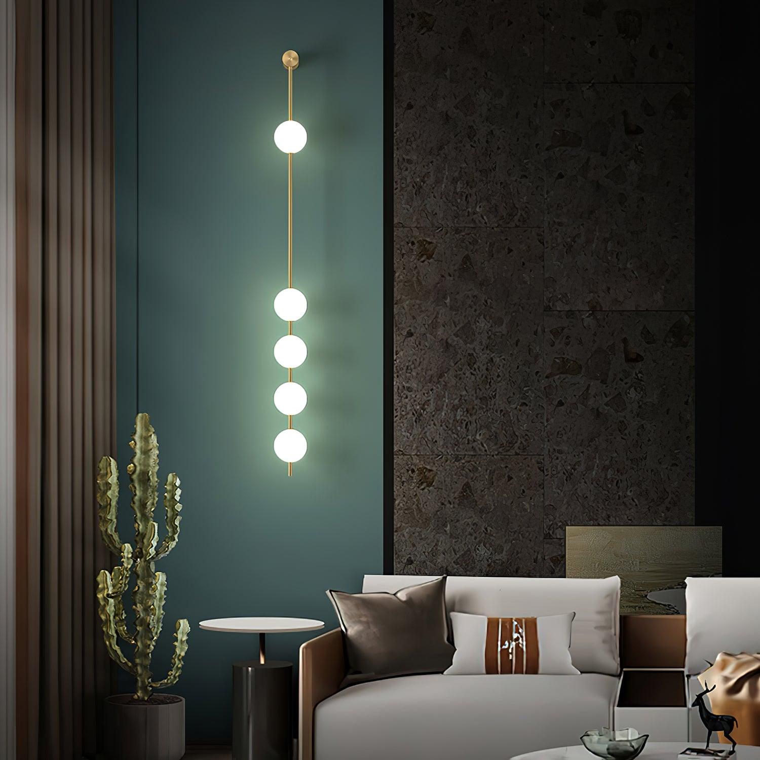 Wall lamp with a vertical row of white ball shades on a slim brass-coloured rod lit against a dark green wall panel.