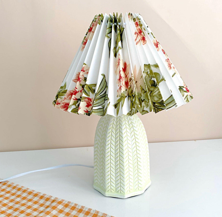 Table lamp with a white pleated shade in a printed floral pattern and a pale green ceramic base with a raised chevron pattern lit on a white table with a checked cloth.