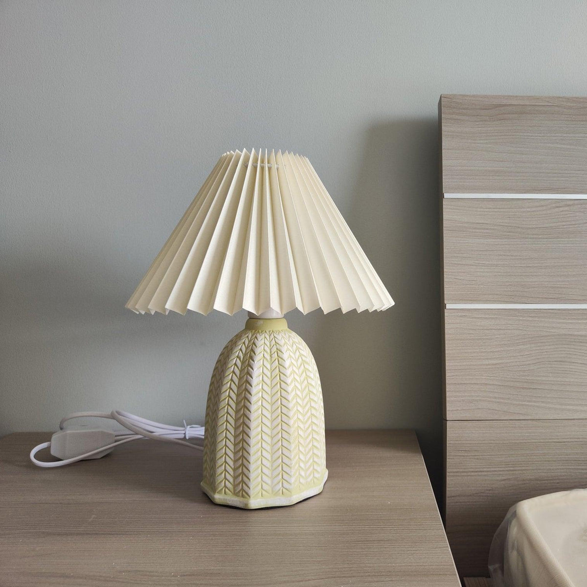 Table lamp with a plain cream pleated shade and a pale green ceramic base with a raised chevron pattern on a bedside table beside a wooden headboard.