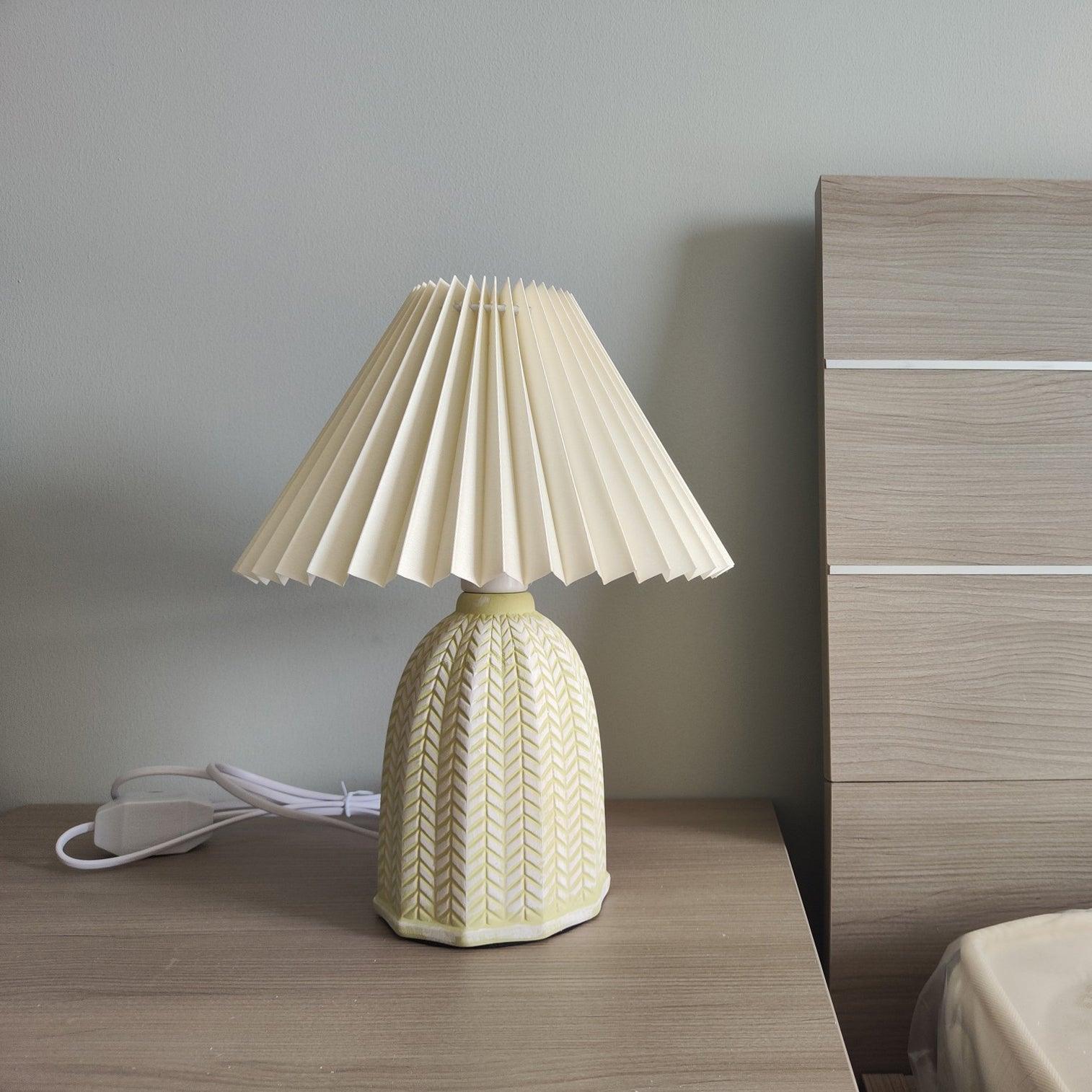 Table lamp with a plain cream pleated shade and a pale green ceramic base with a raised chevron pattern on a bedside table beside a wooden headboard.
