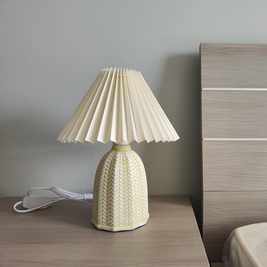 Table lamp with a plain cream pleated shade and a pale green ceramic base with a raised chevron pattern on a bedside table beside a wooden headboard.