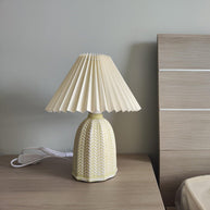 Table lamp with a plain cream pleated shade and a pale green ceramic base with a raised chevron pattern on a bedside table beside a wooden headboard.
