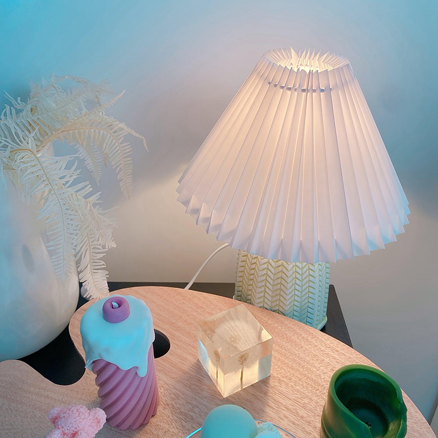 Table lamp with a plain cream pleated shade and a pale green ceramic base with a raised chevron pattern lit on a round table beside small ornaments.