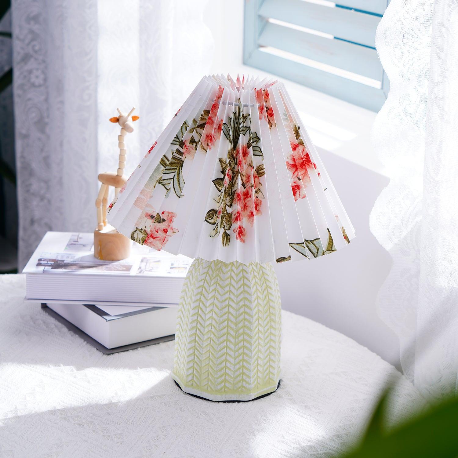 Table lamp with a white pleated shade in a printed floral pattern and a pale green ceramic base with a raised chevron pattern lit on a table beside a wooden figure and magazines.