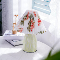 Table lamp with a white pleated shade in a printed floral pattern and a pale green ceramic base with a raised chevron pattern lit on a table beside a wooden figure and magazines.