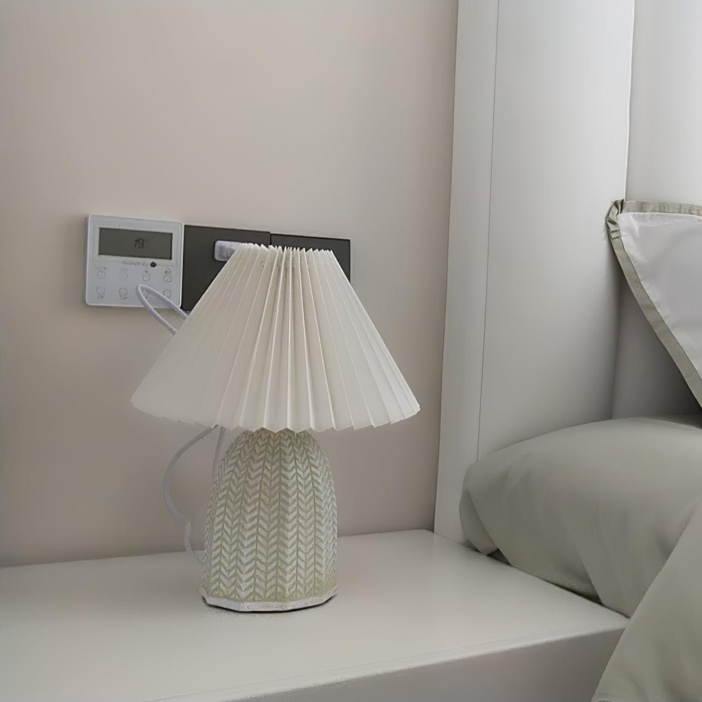 Table lamp with a plain cream pleated shade and a pale green ceramic base with a raised chevron pattern on a bedside table beside a bed.