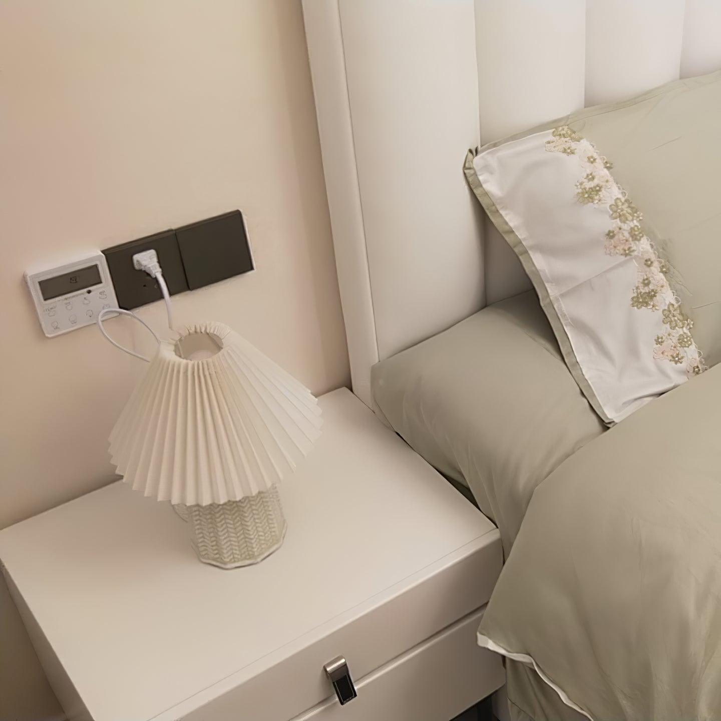 Table lamp with a plain cream pleated shade and a pale green ceramic base with a raised chevron pattern lit on a bedside table beside a bed.
