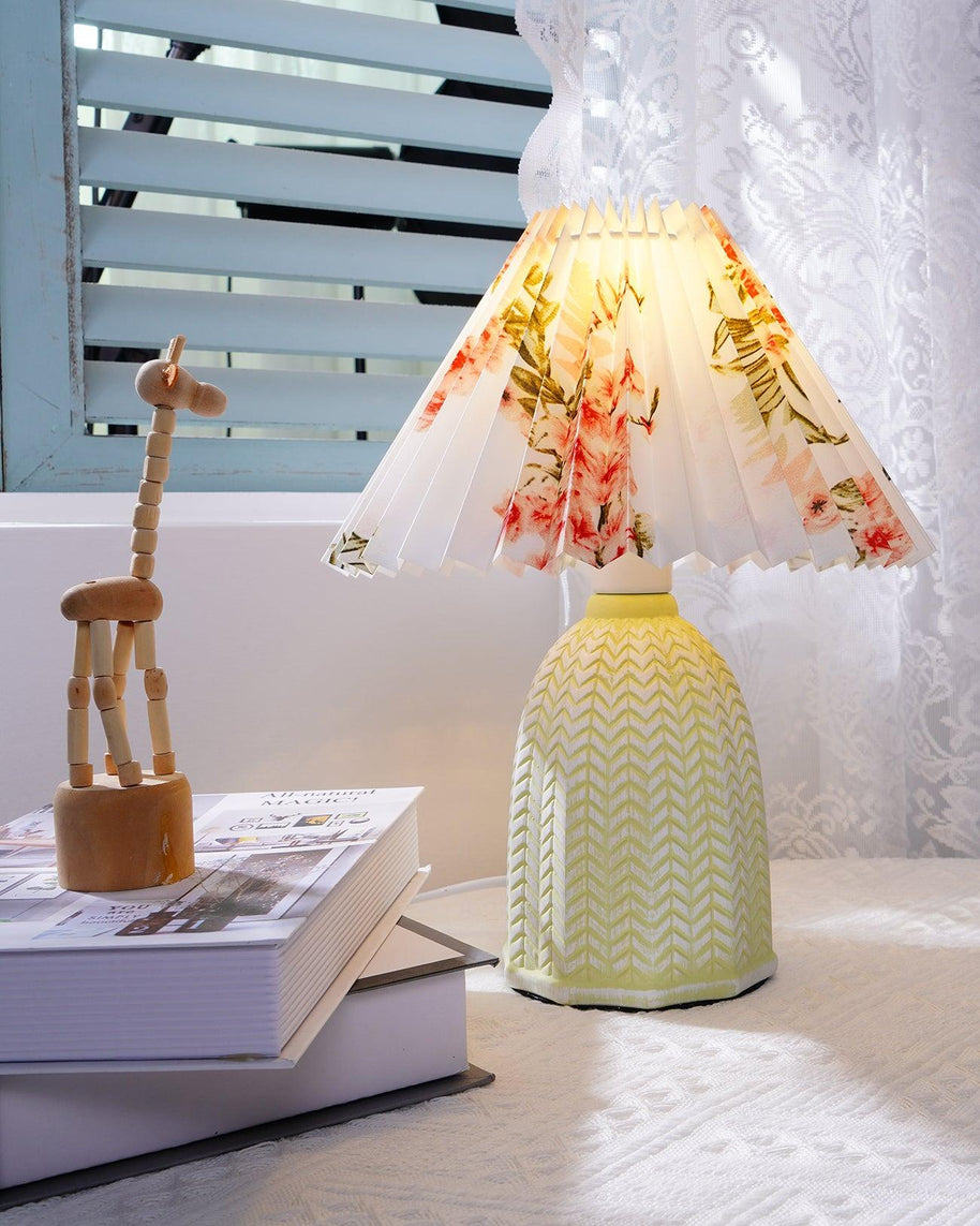 Table lamp with a white pleated shade in a printed floral pattern and a pale green ceramic base with a raised chevron pattern lit on a windowsill beside a wooden figure and a book.