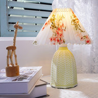 Table lamp with a white pleated shade in a printed floral pattern and a pale green ceramic base with a raised chevron pattern lit on a windowsill beside a wooden figure and a book.
